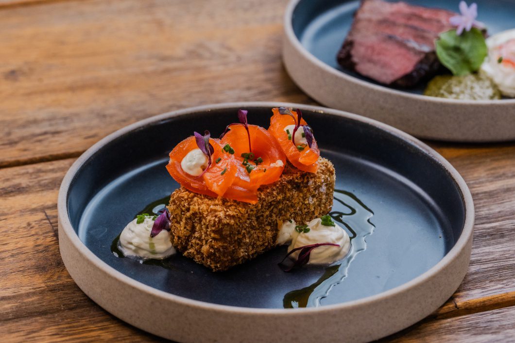 Breaded salmon cube topped with sliced cured salmon and microgreens, served with dollops of sauce on a dark plate.