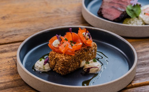 Breaded salmon cube topped with sliced cured salmon and microgreens, served with dollops of sauce on a dark plate.