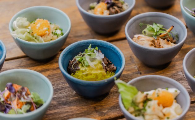 Selection of small plated dishes in colourful ceramic bowls, including seafood, vegetables, and grains on a wooden table.