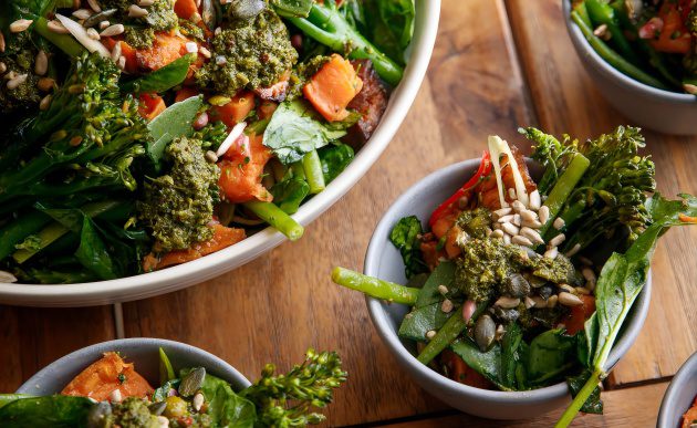 Large serving bowl of leafy green salad with roasted vegetables, dollops of pesto, and a mix of sunflower and pumpkin seeds, surrounded by smaller individual portions on a wooden table.
