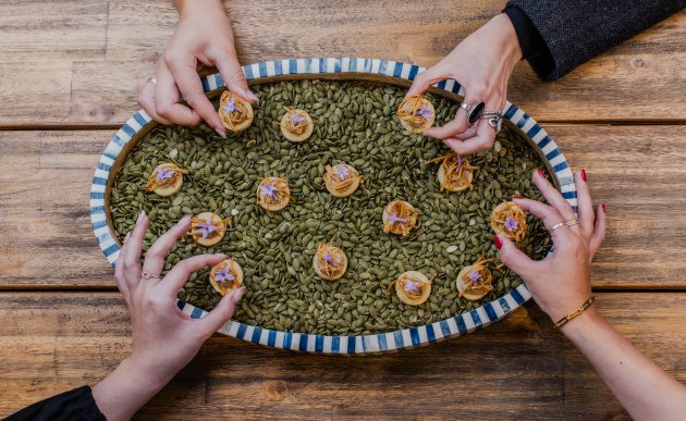 Overhead view of hands reaching for bite-sized canapés arranged on a platter filled with green seeds on a wooden table.