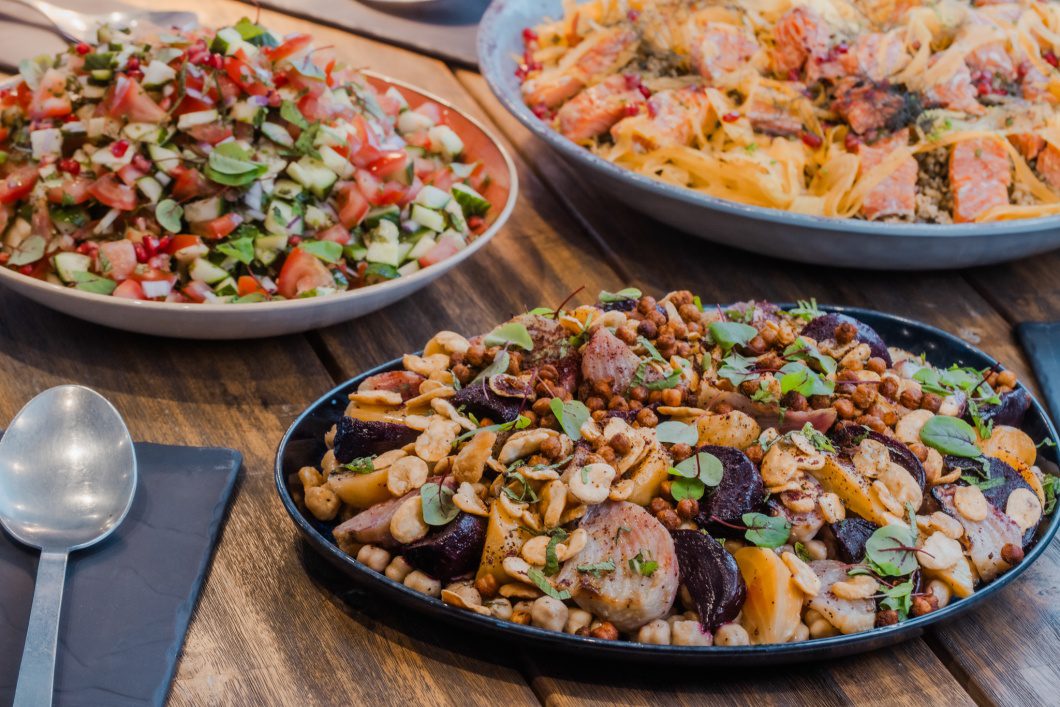 Table spread of vibrant sharing dishes including roasted vegetables with chickpeas and seeds, a fresh chopped salad, and a grain-based dish topped with fish and herbs.