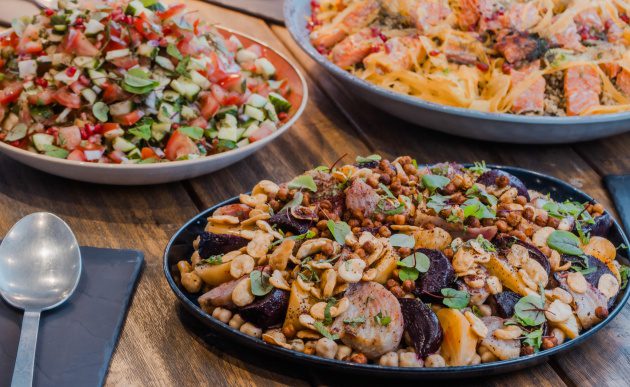 Table spread of vibrant sharing dishes including roasted vegetables with chickpeas and seeds, a fresh chopped salad, and a grain-based dish topped with fish and herbs.