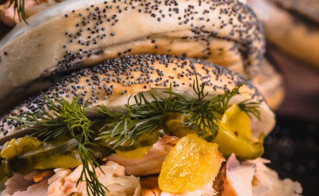 Close-up of stacked poppy seed bagels filled with smoked salmon, cream cheese, pickles, and fresh dill on a serving tray.